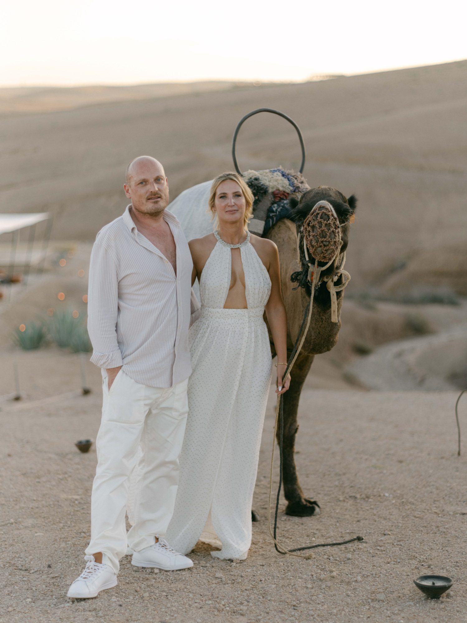 Agafay desert couple camel portrait Couple dressed in white standing beside a decorated camel in the Agafay desert